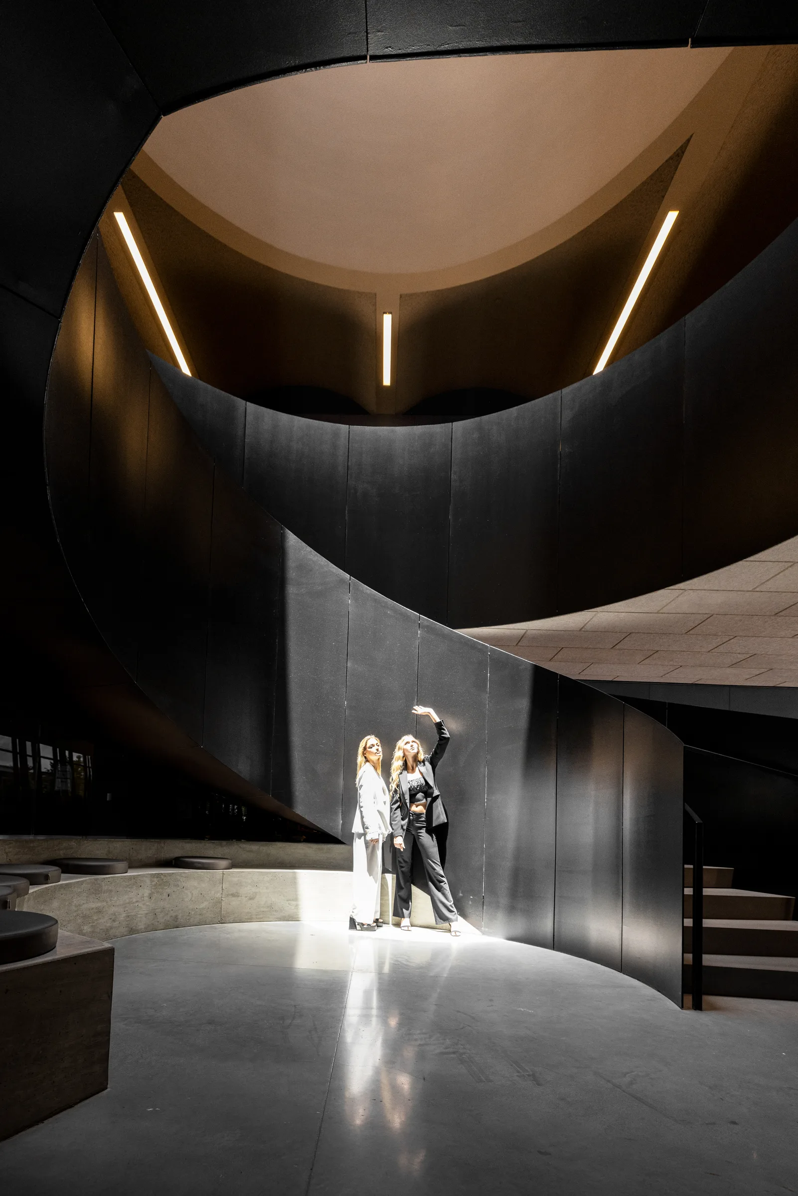 Two women in black and white suits standing beneath a curving black spiral staircase, warm lighting, by ARCHTRAITS