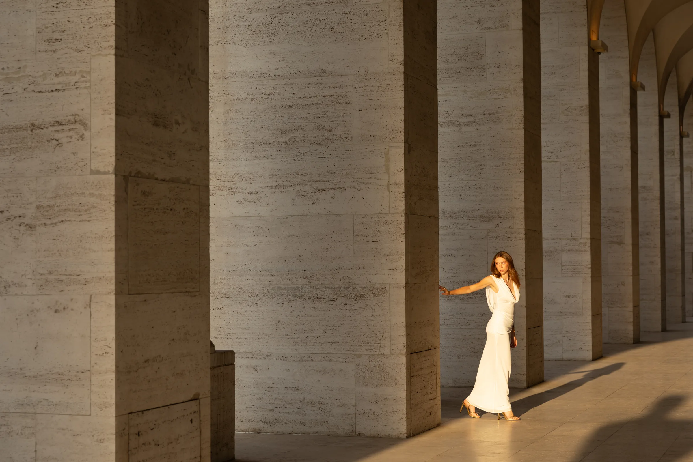 Woman in a white dress walking through a sunlit stone colonnade at Palazzo della Civiltà Italiana, Rome, by ARCHTRAITS