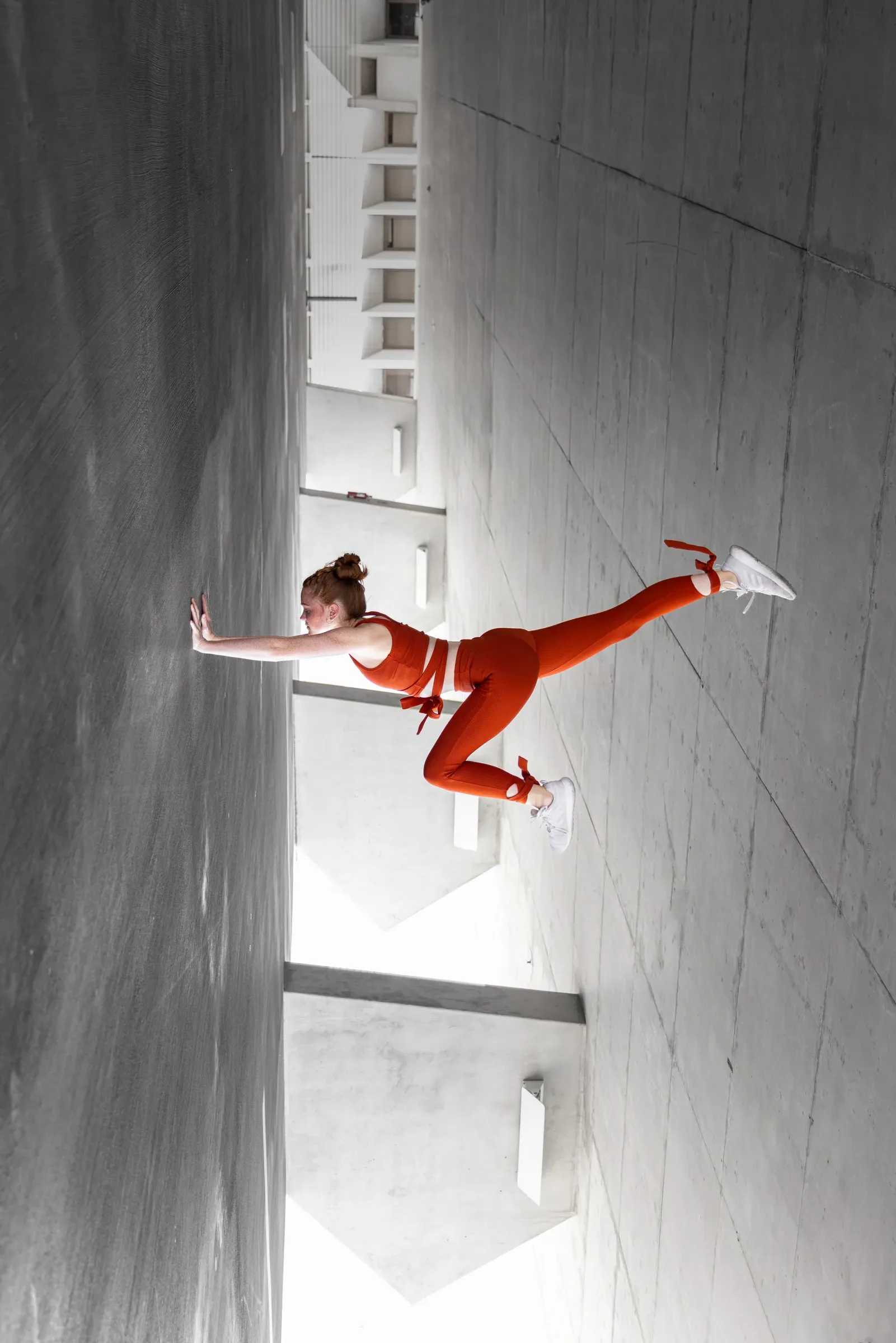 Woman in an orange outfit performing a handstand in a concrete parking garage, by ARCHTRAITS