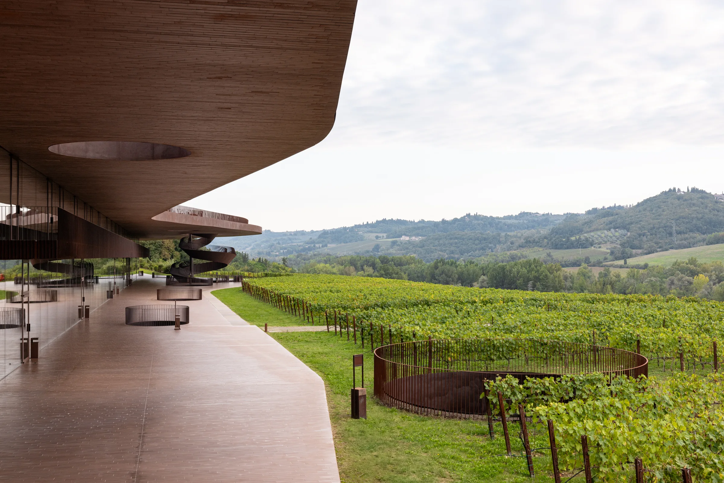 Cantina Antinori's curved bronze roof and spiral staircase seen from the terrace overlooking the vineyard, Tuscany, by ARCHTRAITS