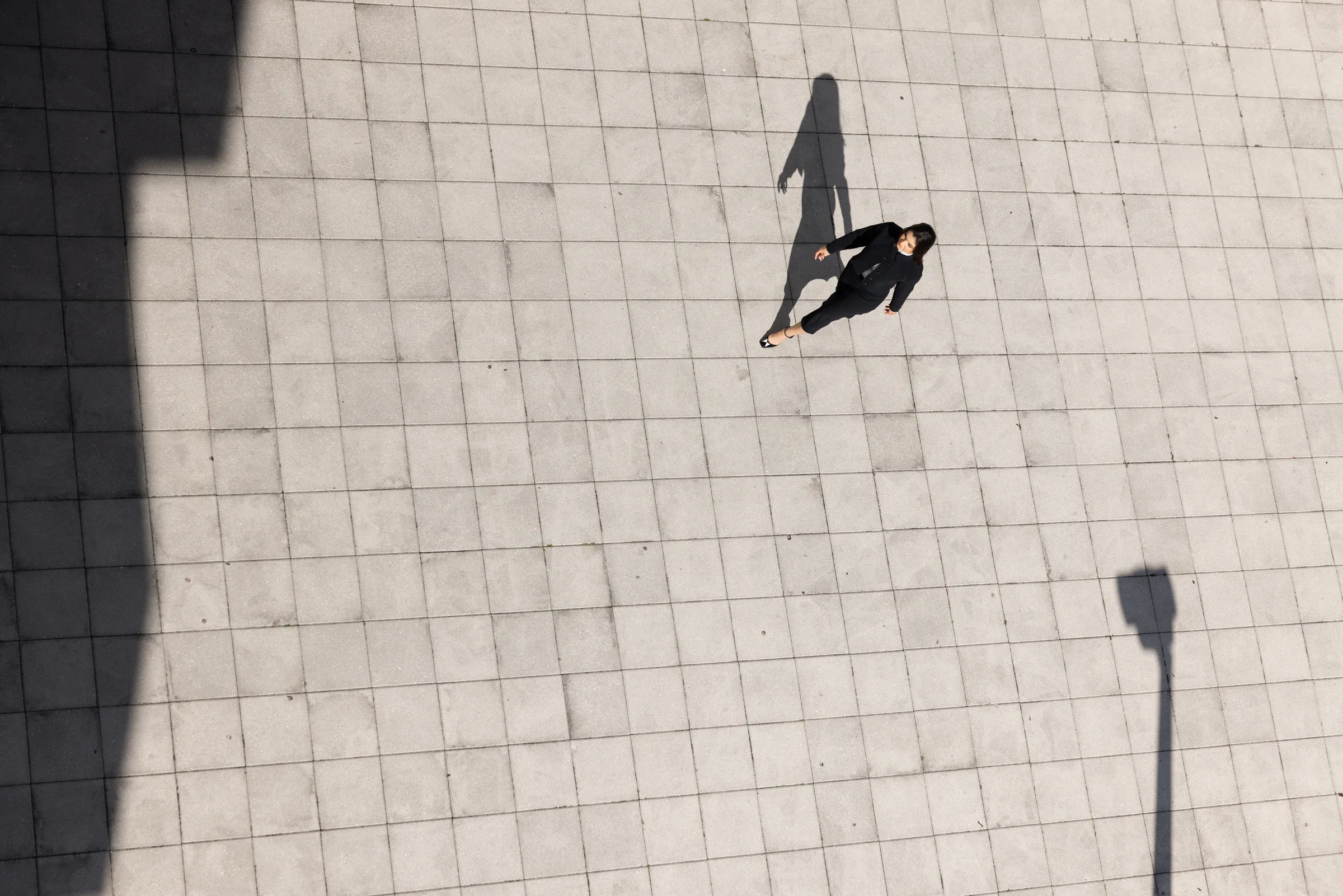 Aerial view of a woman walking across a paved plaza with a long dramatic shadow at Miami Dade College, Florida, by ARCHTRAITS