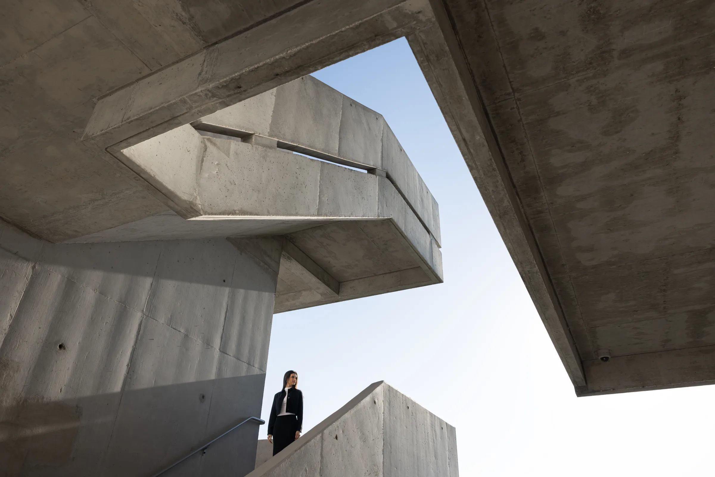 Woman standing at the edge of an angular concrete stairwell landing, looking up through a geometric cutout, Miami Dade College, Florida, by ARCHTRAITS