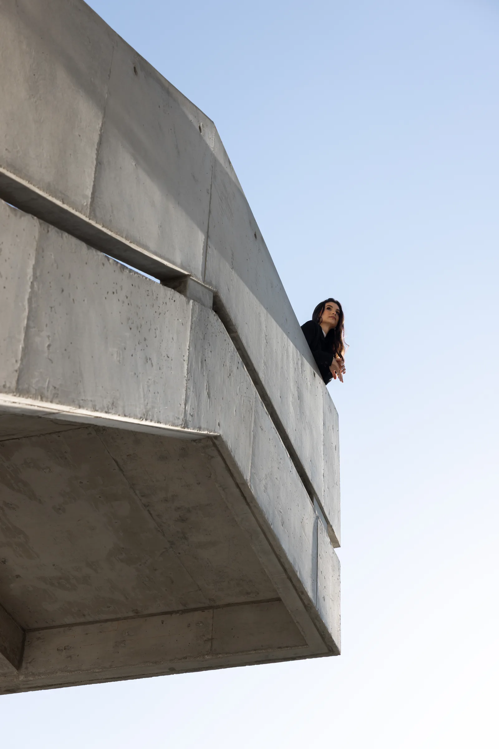 Woman leaning over the edge of a cantilevered concrete balcony against a clear blue sky at Miami Dade College, Florida, by ARCHTRAITS