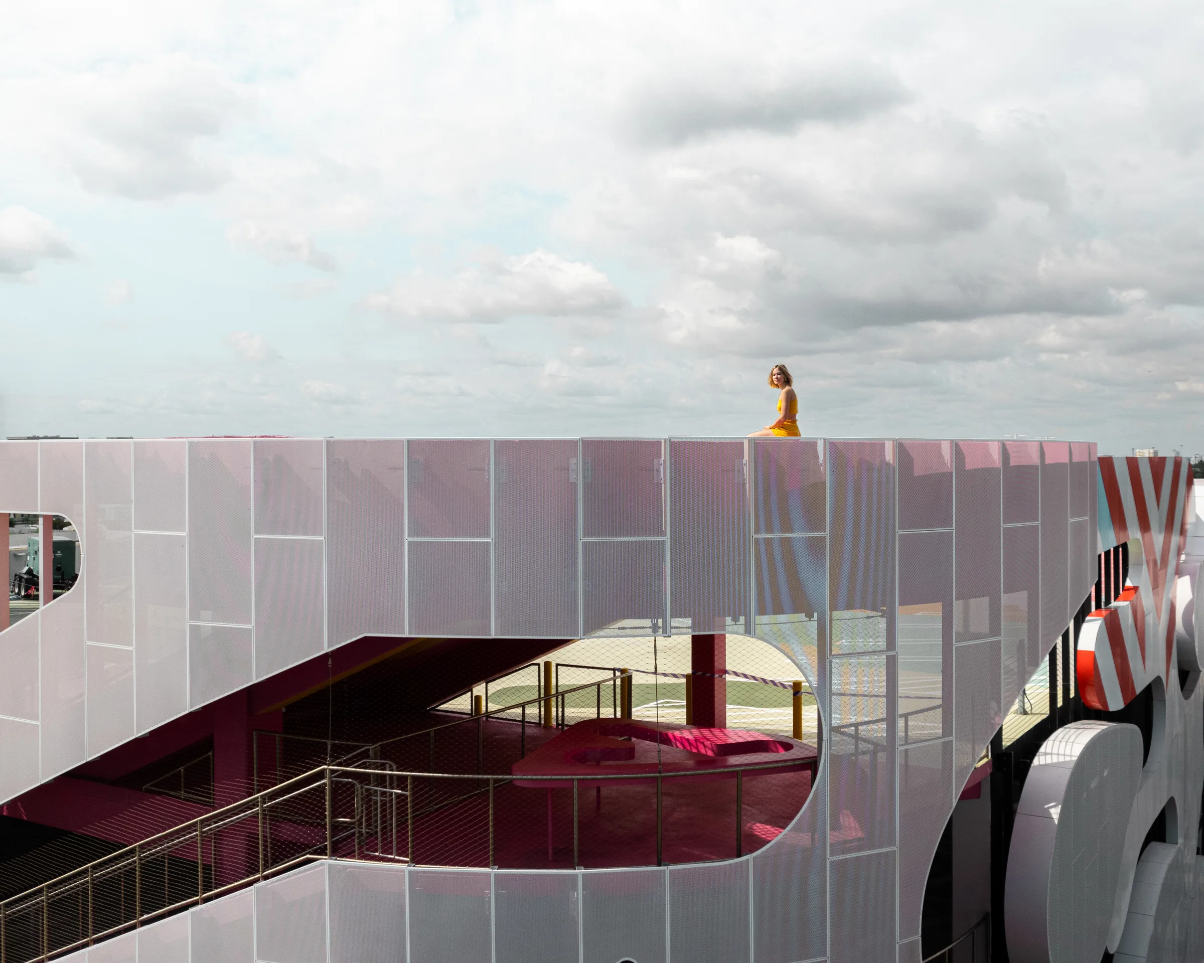 Woman in yellow sitting atop Museum Garage's perforated white parapet, overlooking its red-and-white striped rooftop, Miami Design District, by ARCHTRAITS