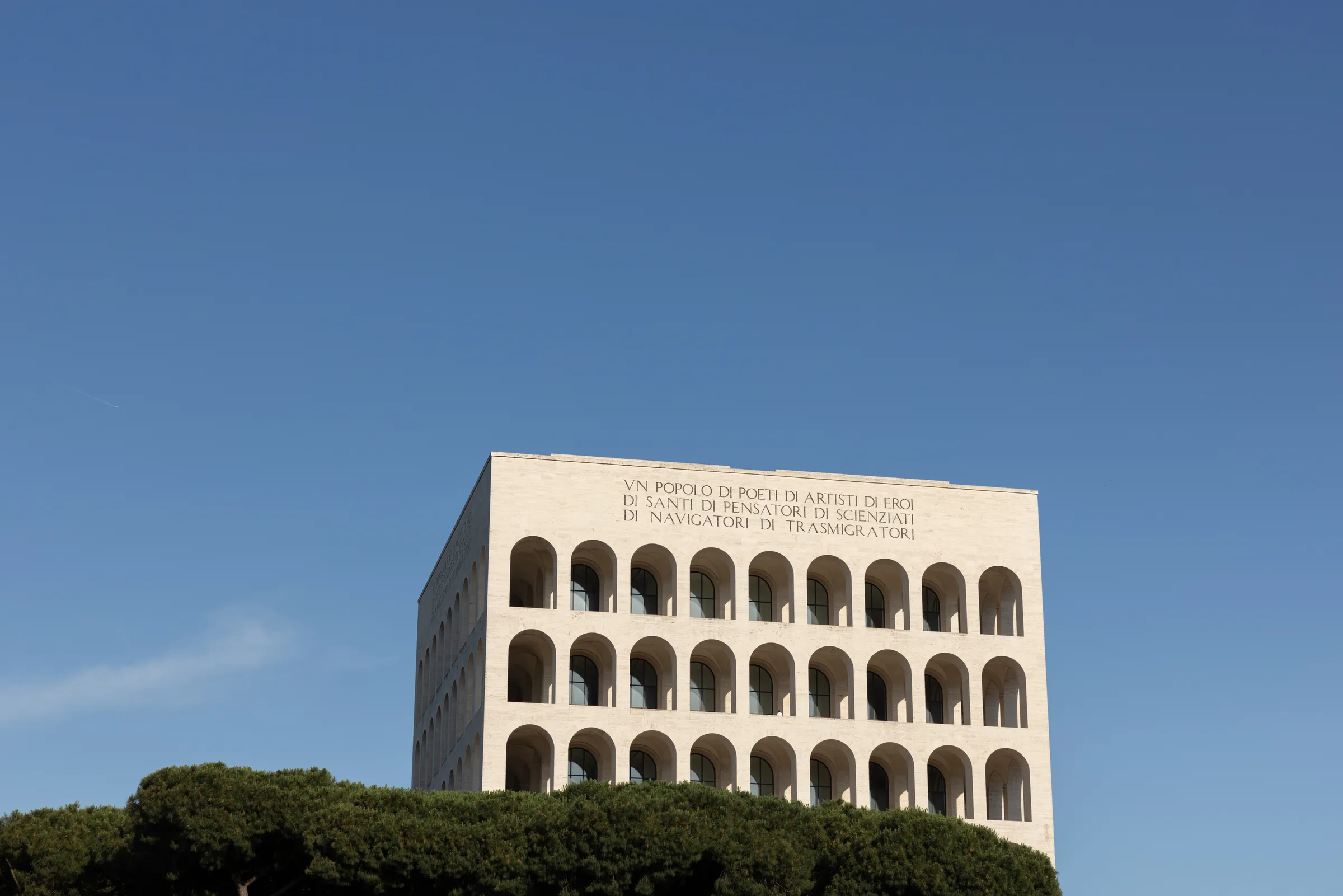 Palazzo della Civiltà Italiana's travertine facade with its inscribed motto, rising above pine trees against a clear sky, Rome, by ARCHTRAITS