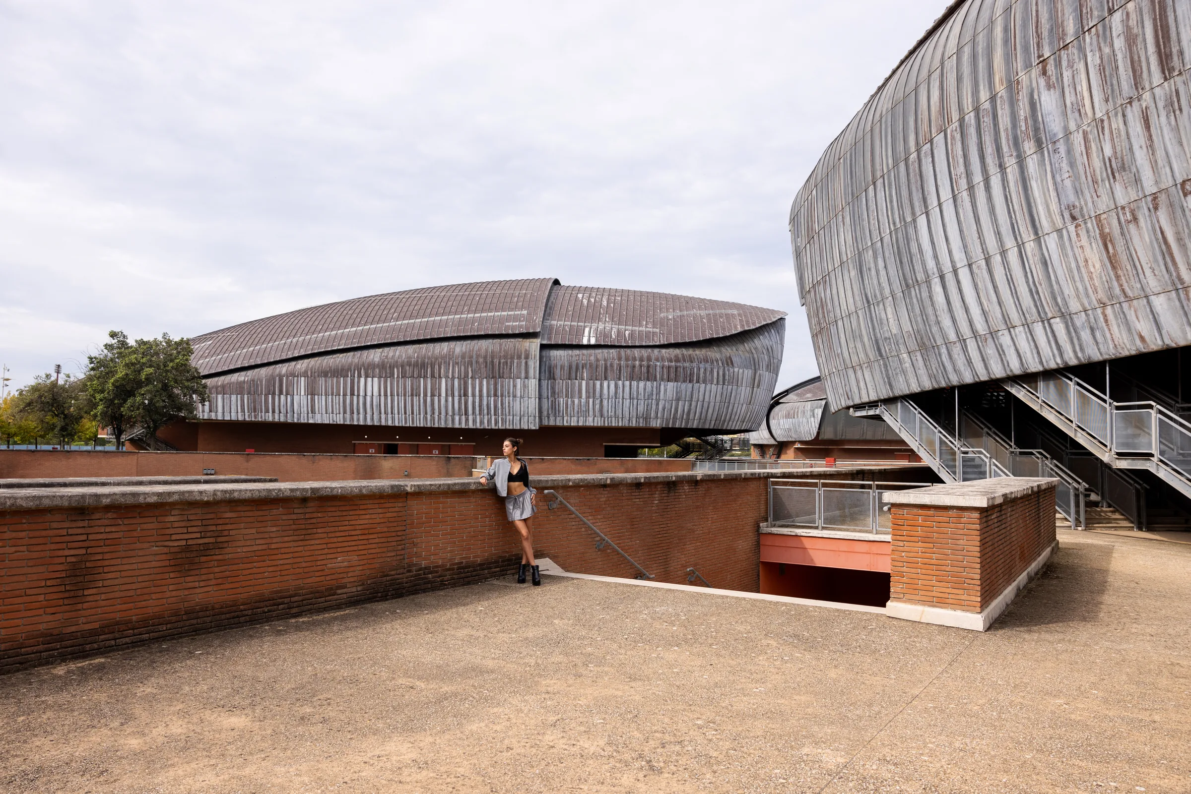 Woman leaning against a brick wall overlooking Auditorium Parco della Musica's zinc-clad concert hall shells, Rome, by ARCHTRAITS