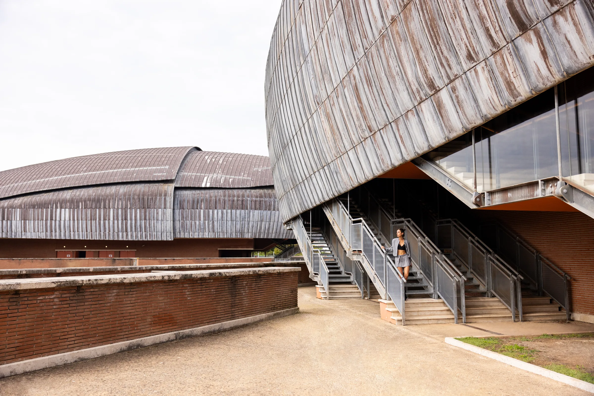 Woman descending a staircase between Auditorium Parco della Musica's zinc-clad concert hall shells, Rome, by ARCHTRAITS